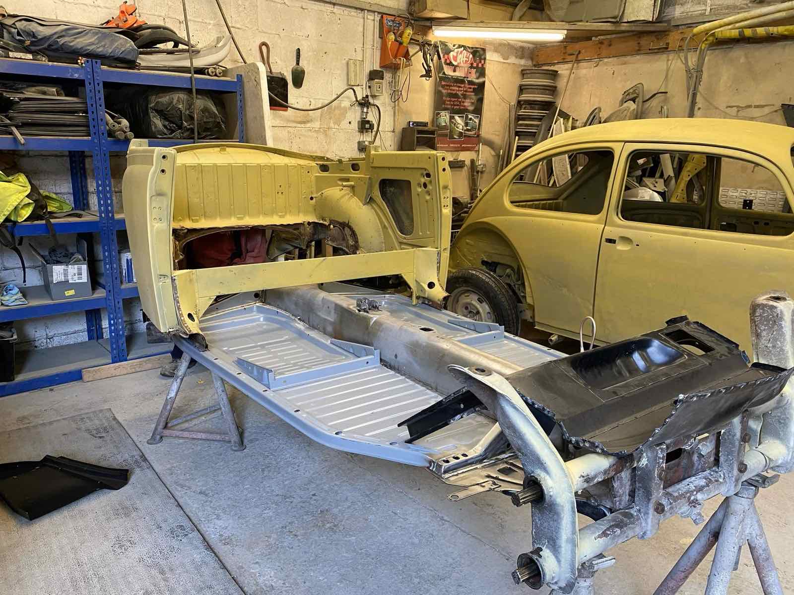 A Beetle floor pan and body tub on stands in the workshop, another Beetle shell behind