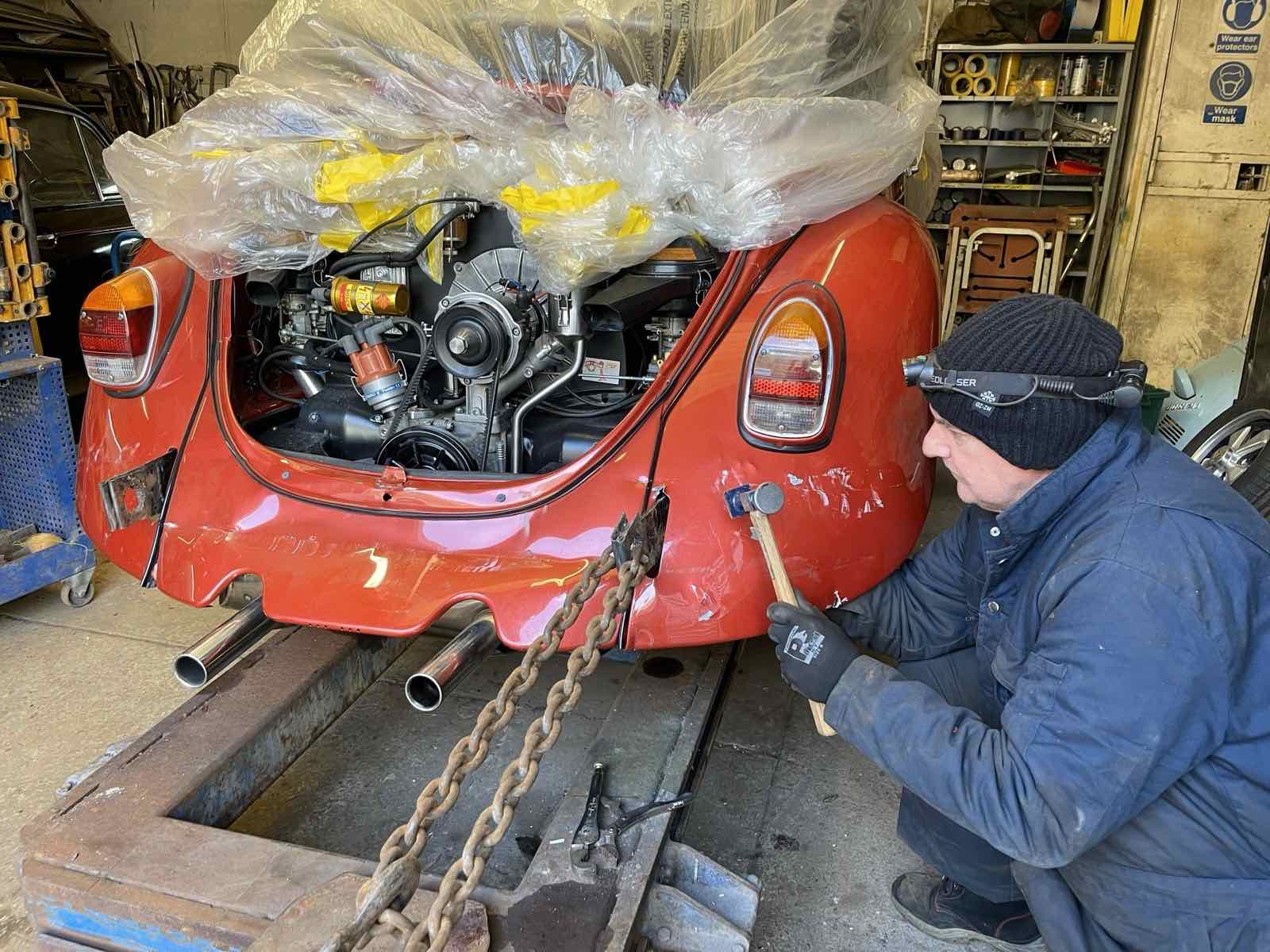 A mechanic in a head torch working on the rear of a red Beetle cabriolet, engine bay open