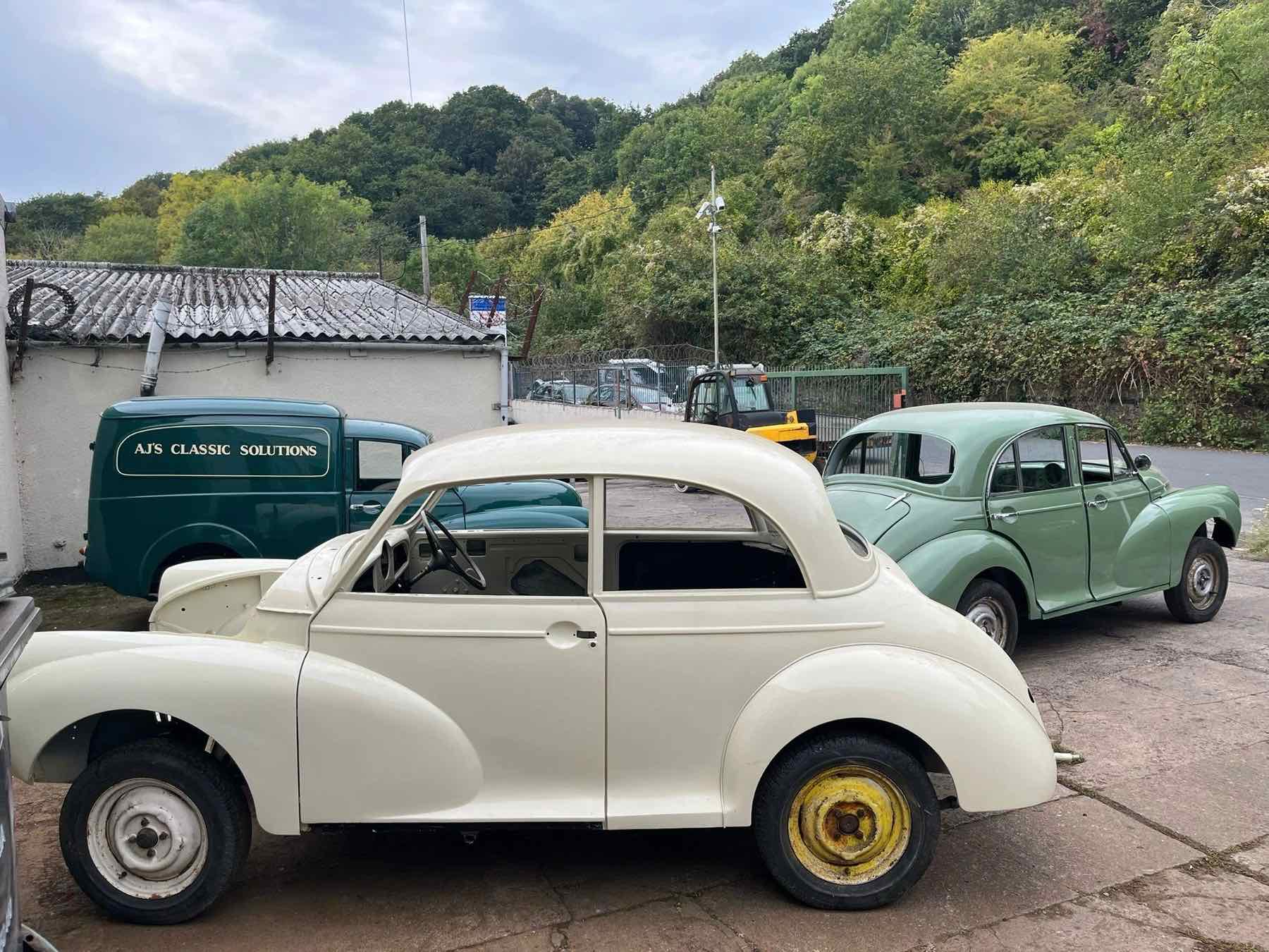 A finished green Morris Minor saloon in the yard, with a cream Morris shell and the wooded hillside behind
