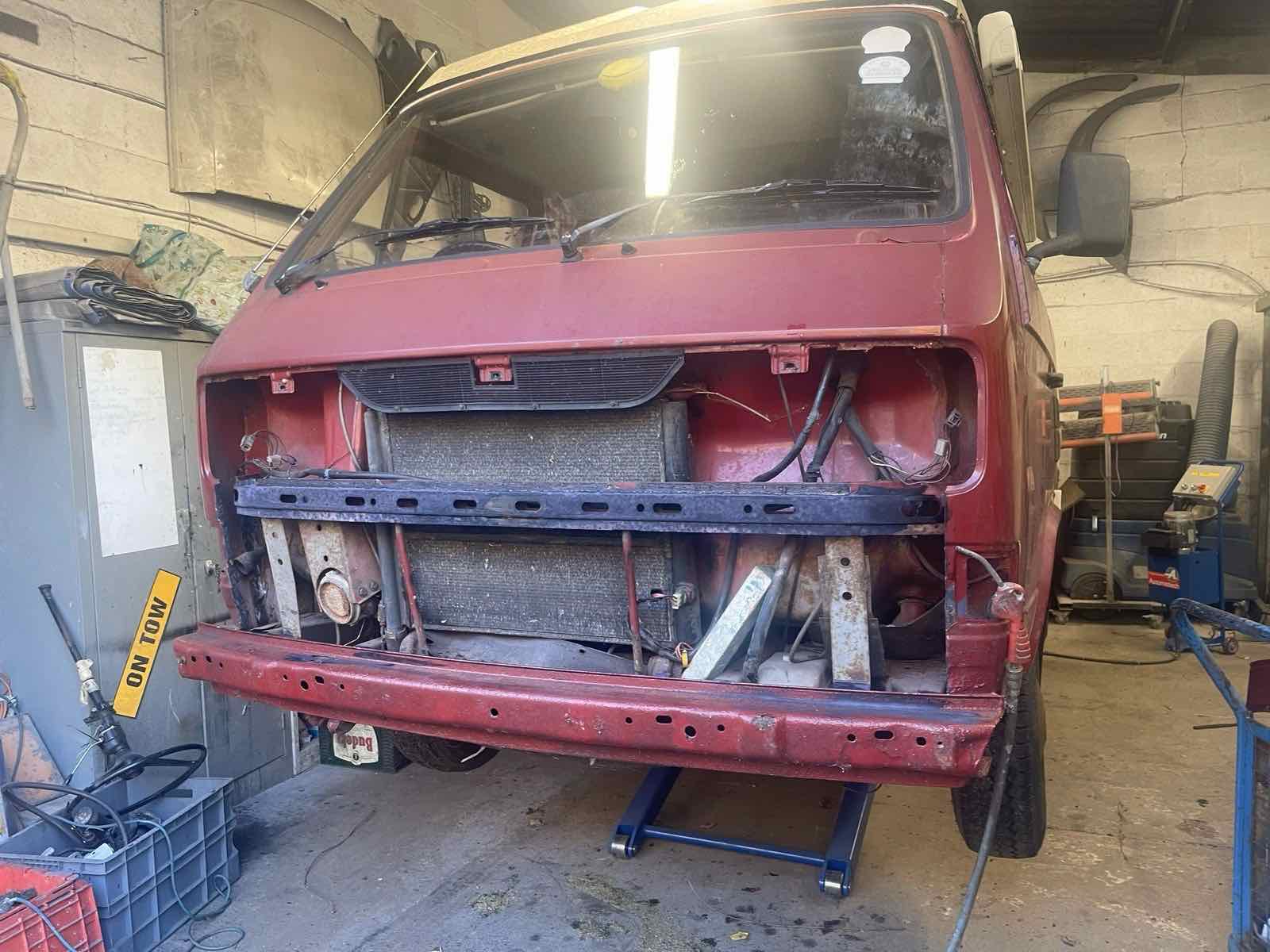 A red VW T25 camper with its front end stripped back in the workshop